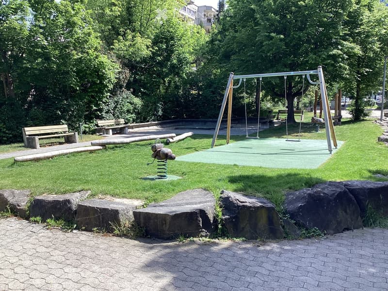 A park area with a swing set, benches, a spring-loaded toy, and large rocks arranged on a paved surface with grassy areas.