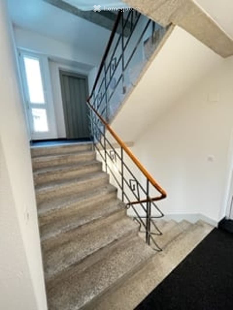 A modern staircase with a wooden handrail and metal balustrade, leading up to a higher level. The walls are painted white, and the carpeted steps are visible.