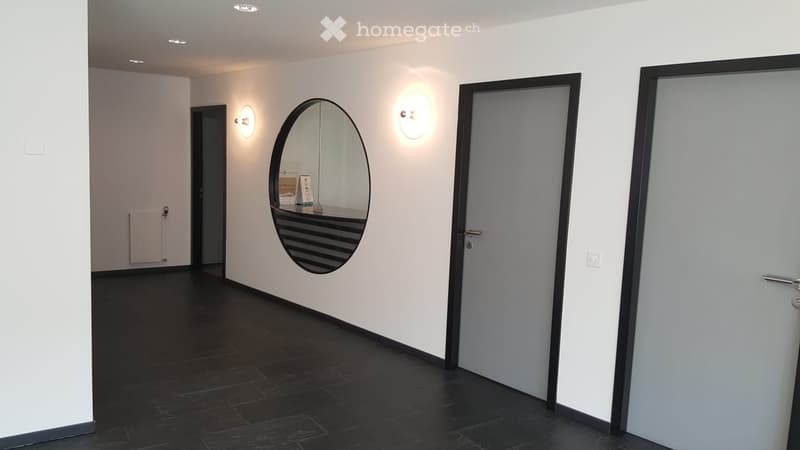 Modern hallway with dark tiled flooring, white walls, and recessed lighting. Features an oval mirror on one wall and two doors, one leading to a room with a visible radiator.