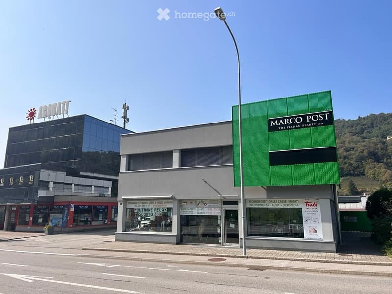 Modern commercial building with glass facade, signage for "MARCO POST THE ITALIAN BEAUTY SPA" and "POLTRONE RELAX SCONTO 50%", located on a paved street with a clear blue sky and distant hills.