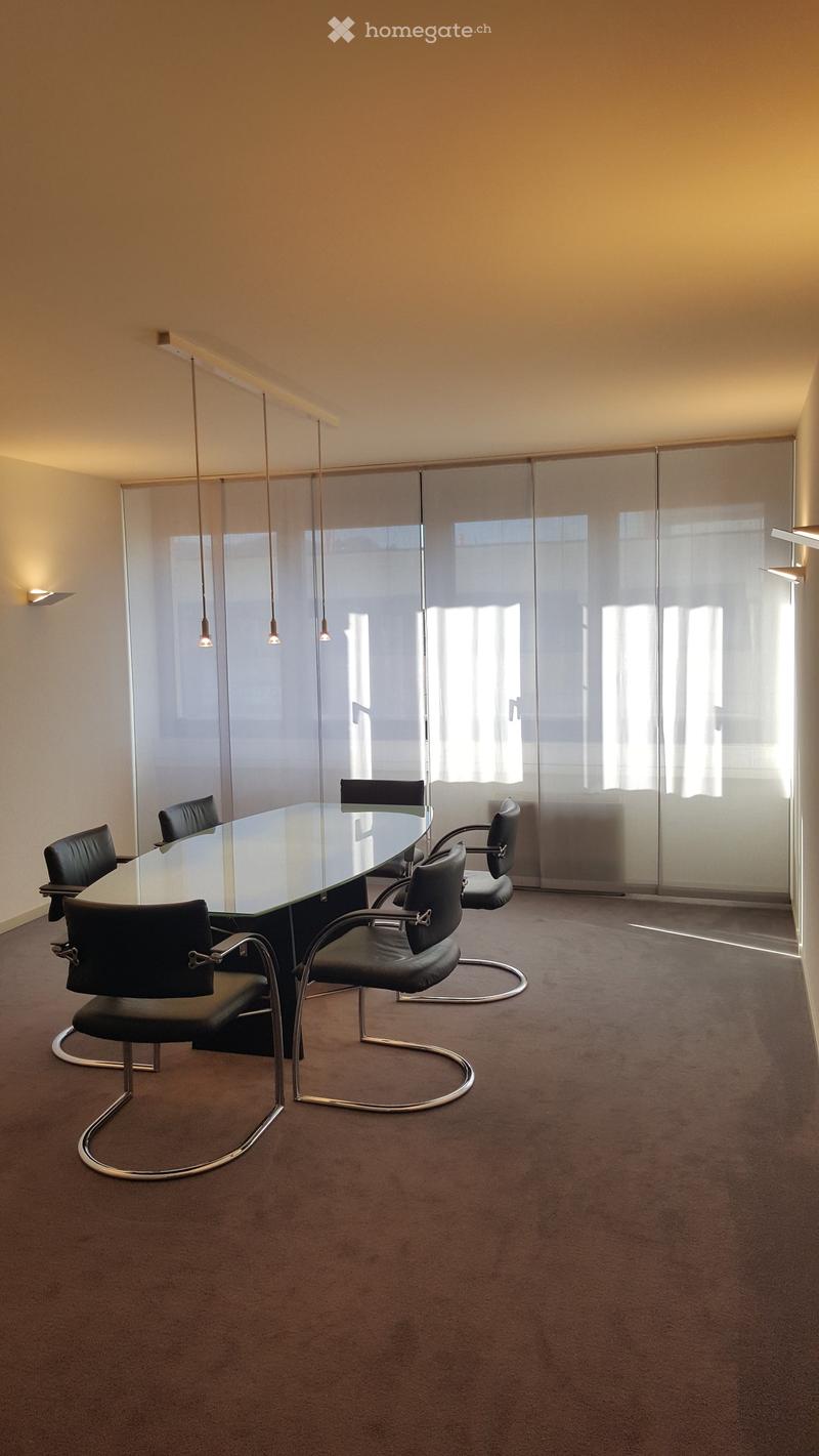Modern conference room with a large glass table surrounded by black leather chairs, pendant lights, and a large glass partition wall.