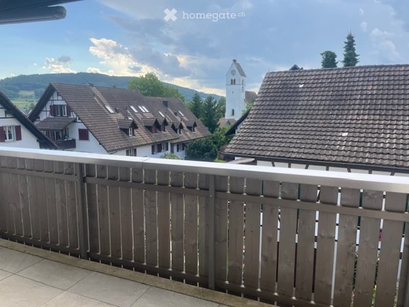 Balcony overlooking a village with traditional houses and a church tower in the background, under a partly cloudy sky.