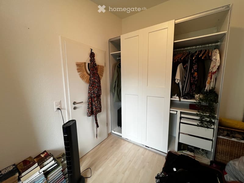 A small, well-lit room with light wooden flooring and white walls. Features include a tall black fan, a stack of books, an open wardrobe with clothes and storage drawers, and a hanging decorative item on the wall.