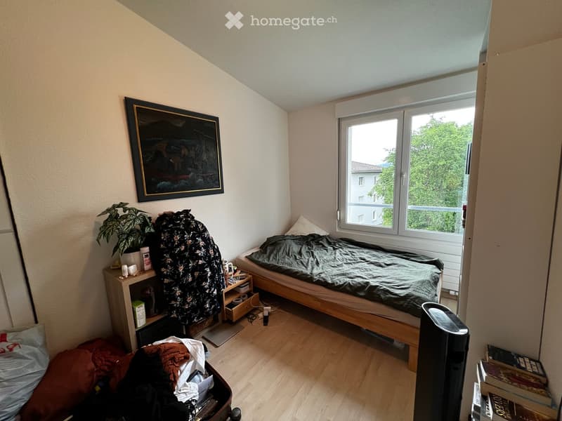 Cozy bedroom with a single bed, nightstand, plant, picture frame, suitcase, and books on the floor.