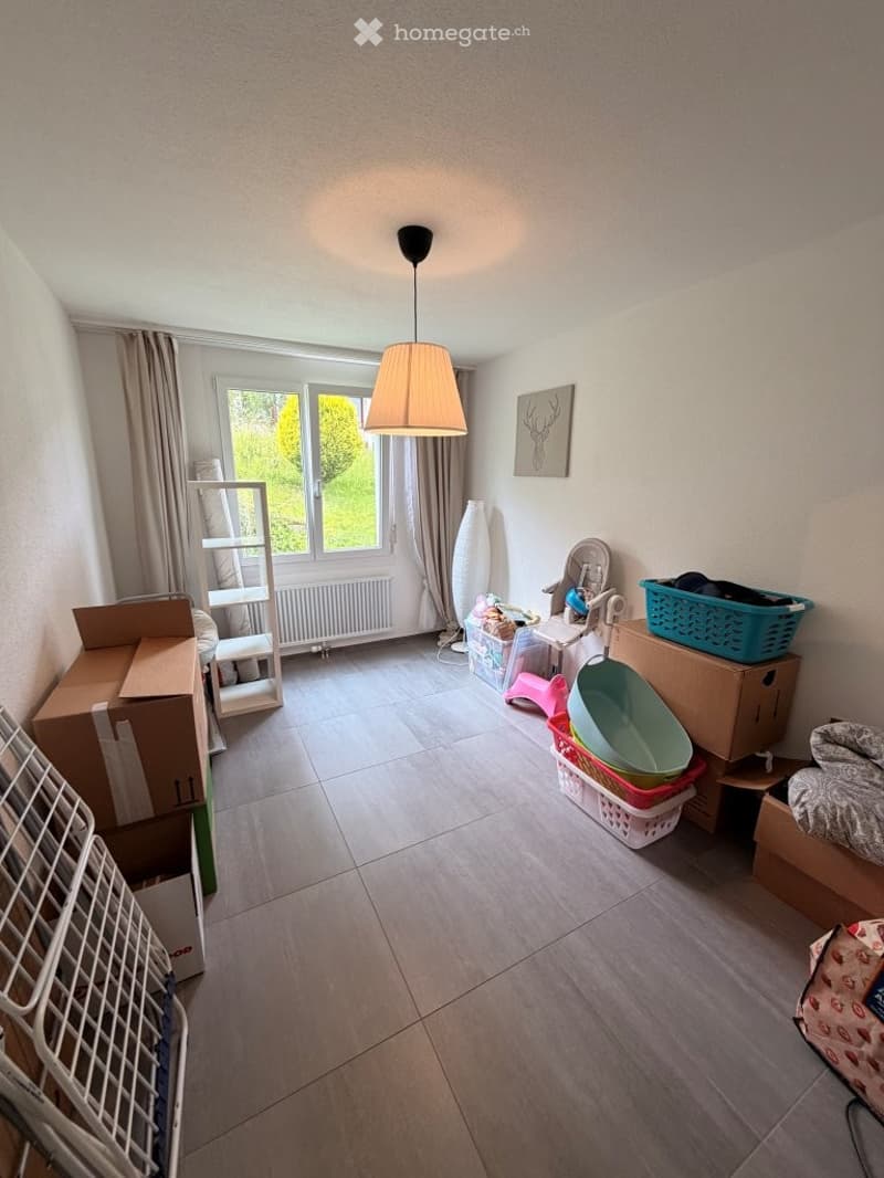 A room with tiled flooring, a large window with curtains, a hanging lamp, a baby chair, a baby bath, storage boxes, a shelf, a radiator, and a picture frame on the wall.