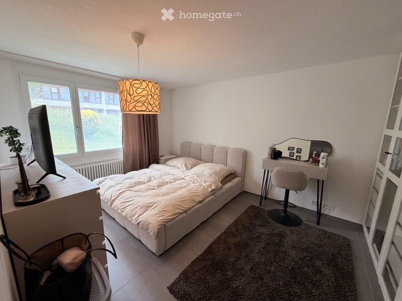 A modern bedroom with a large bed, a vanity table with a mirror, a chair, a screen, a plant, and a hanging lamp.