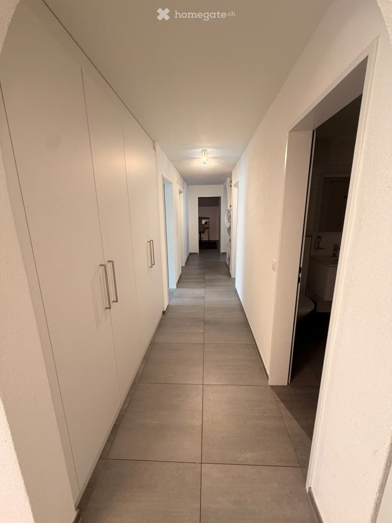 A long, narrow hallway with white walls, tiled floor, and built-in white cabinets on one side. The hallway leads to a room with a washing machine and a bathroom with a sink and toilet.