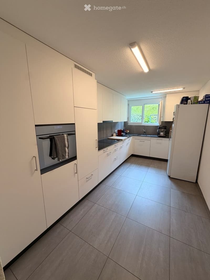Modern kitchen with white cabinets, stainless steel appliances, and grey tiled flooring. Features include a refrigerator, microwave, oven, sink, and coffee maker.