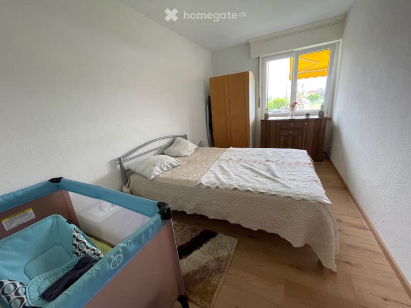 A bedroom featuring a double bed, white bedding, a bedside table, a rug, a wardrobe, a chest of drawers, and a crib.