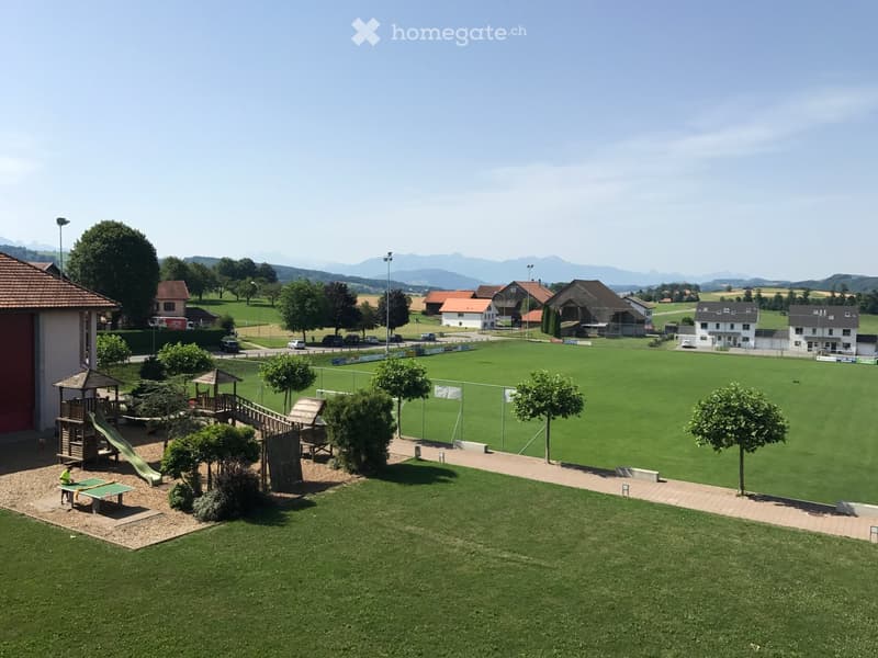 Well-maintained garden with trees, play equipment, table tennis, and a large grassy field. In the distance, houses and mountains are visible.
