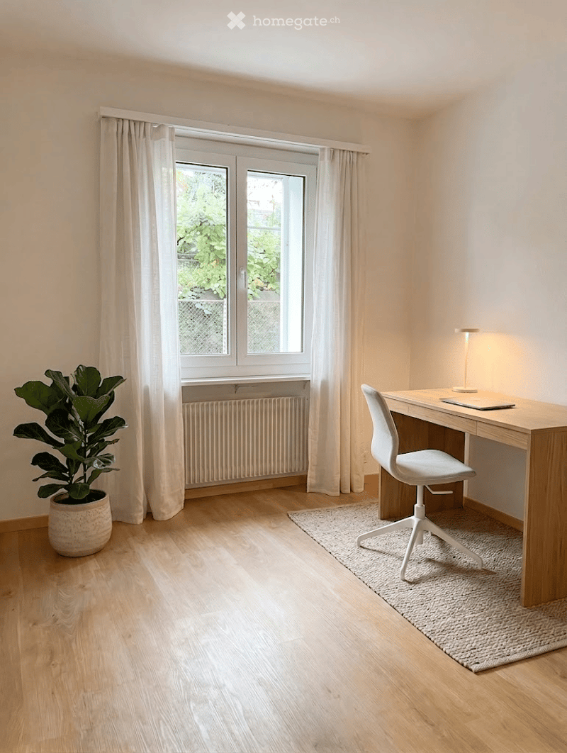 Bright, minimalist room with wooden flooring, large window with white curtains, a modern desk with a chair, a laptop, a small lamp, and a potted plant.