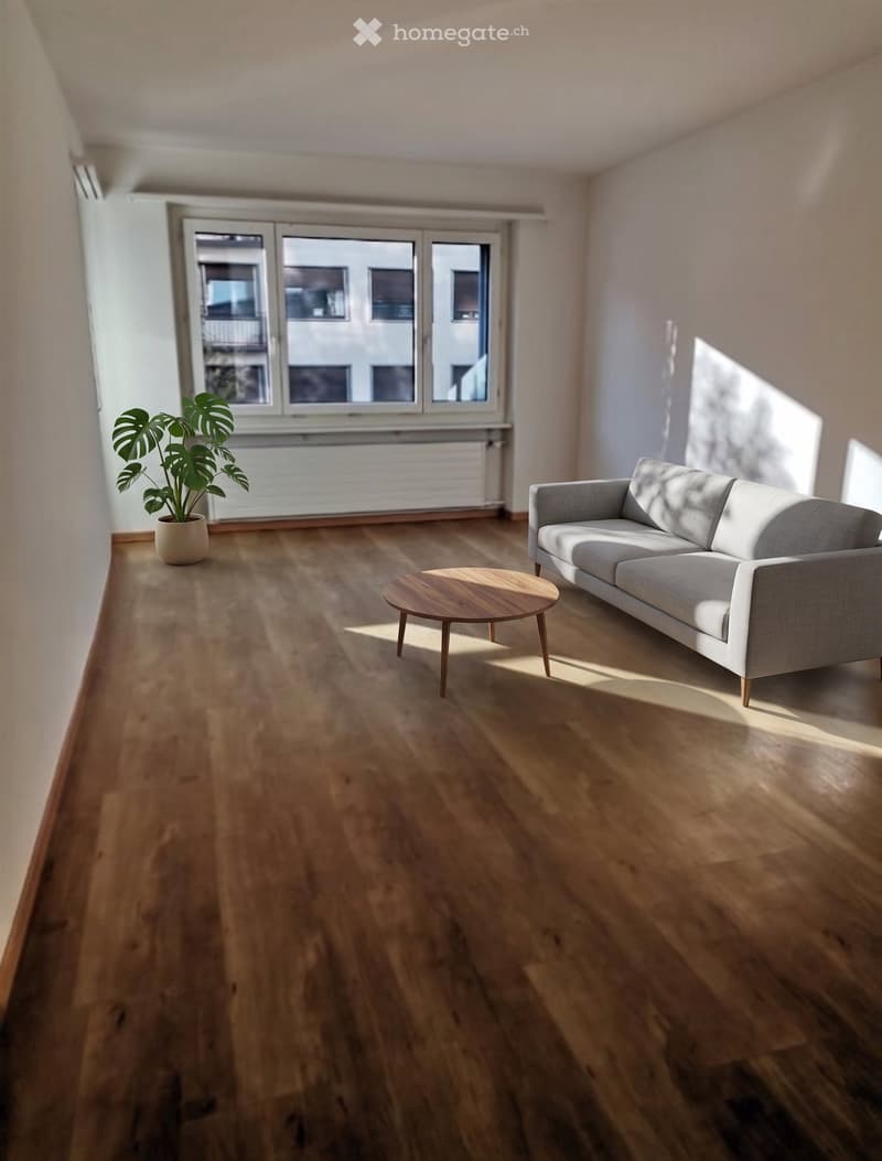 Modern living room with light wooden flooring, a grey sofa, a round wooden coffee table, and a potted plant near the window.