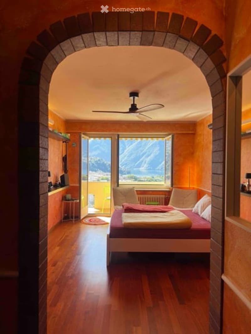 Bright bedroom with wooden floors, large window offering a scenic mountain view, bed with pillows and blanket, nightstand, small table, and a ceiling fan.