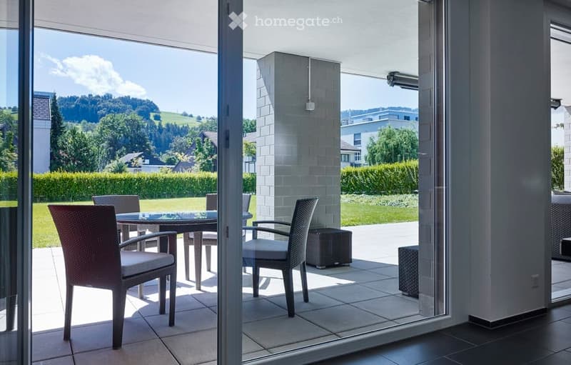 Balcony with a glass table and four wicker chairs, overlooking a green landscape with trees and houses.