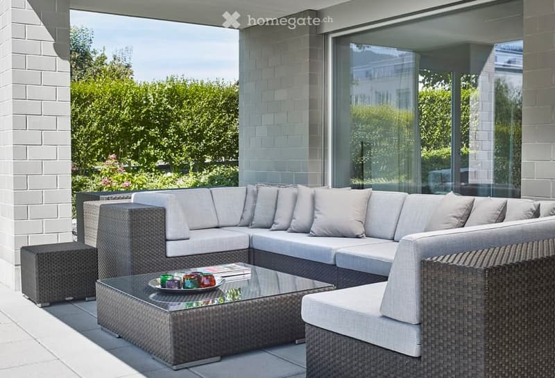 Modern outdoor seating area with a grey sectional sofa, glass coffee table, and matching stool, set against a backdrop of lush greenery.