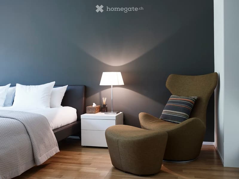 Modern bedroom with a dark grey wall, a wooden floor, a bed with white bedding, a nightstand with a lamp, and a green armchair with an ottoman.
