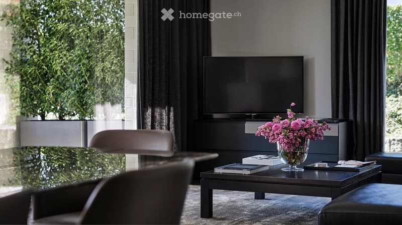 Modern living room with large windows, dark wood furniture, a flat-screen TV, and a glass coffee table with a vase of pink flowers.