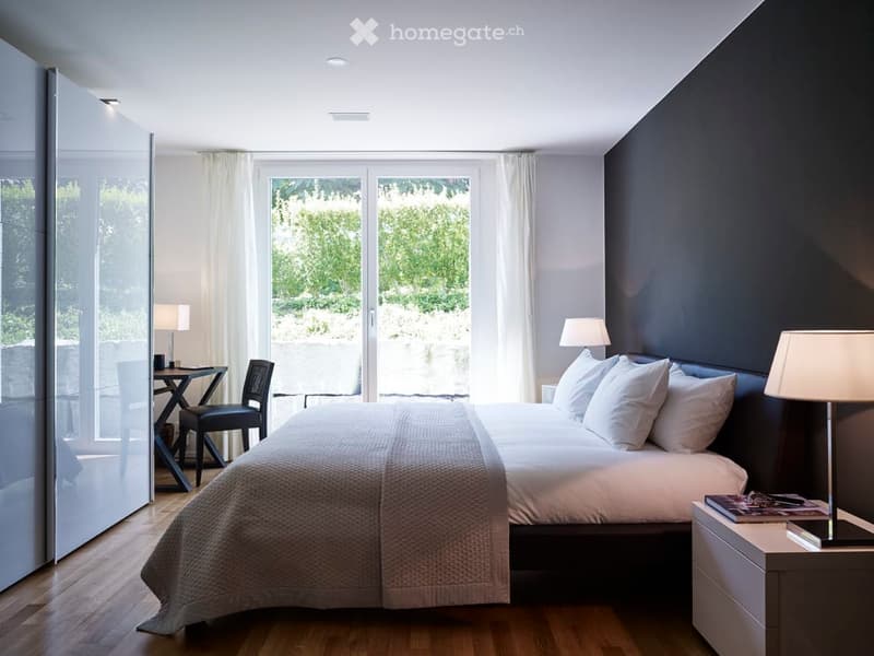 Modern bedroom with a large bed, nightstand, lamp, chair, small table, and large windows with curtains offering a view of greenery.