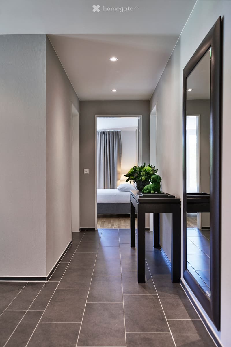 Modern hallway with dark tiled floor, grey walls, and recessed lighting. A small black console table with a vase of greenery and a decorative green animal figurine is placed against the wall. A bedroom is visible at the end of the hallway, featuring a bed