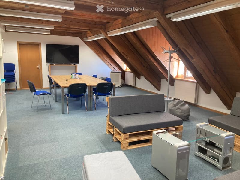 A modern, multi-purpose room with a wooden table, blue chairs, a grey sofa, a TV screen, and computer equipment.