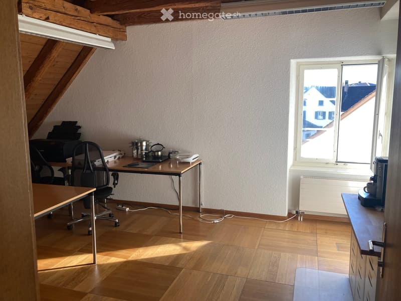 Bright attic room with wooden flooring, slanted wooden ceiling beams, and large window offering a view of rooftops. Contains a desk with a chair, printer, teapot, and other small items, plus a small cabinet with a cup on top.