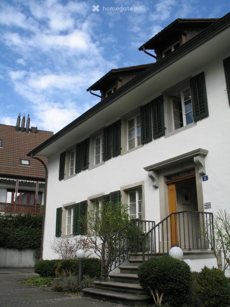 Two-story white house with green shutters, a balcony, and a staircase leading to the entrance.