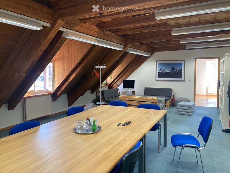 A modern, well-lit room with wooden beams and slanted ceilings. It features a long wooden table surrounded by blue chairs, a small sofa, a screen on a stand, and a picture frame on the wall. The room has large windows and a cozy, inviting atmosphere.