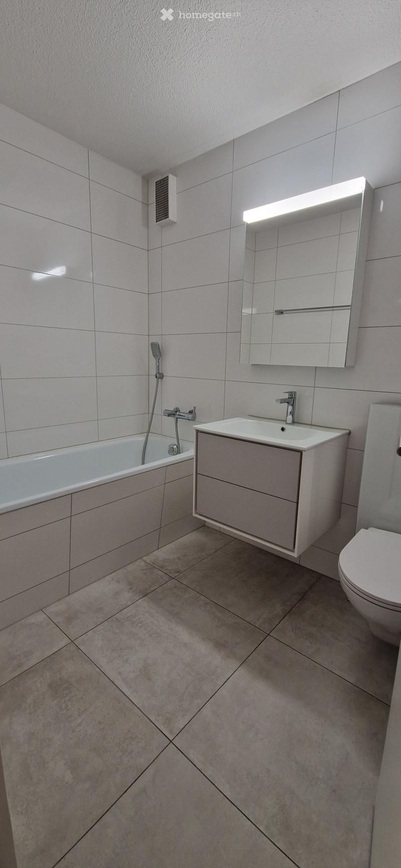 The image shows a modern bathroom with white tiled walls and floors. It has a bathtub, a vanity unit with a sink, and a mirror with a light fixture above it. The bathroom appears to be clean and well-maintained.