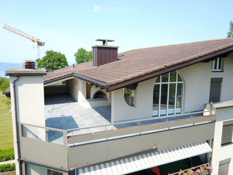 A modern two-story house with a tiled roof, large windows, and a spacious balcony area. The balcony features a wooden railing, a paved floor, and a built-in barbecue area.