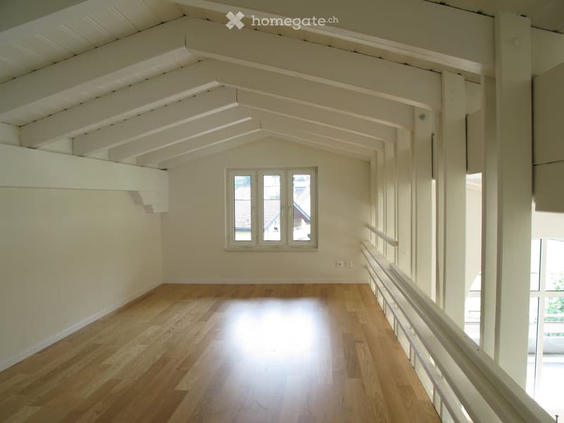 Empty room with wooden flooring, white walls, and a large window with three panes. Exposed wooden beams on the ceiling and a white railing leading to another area.
