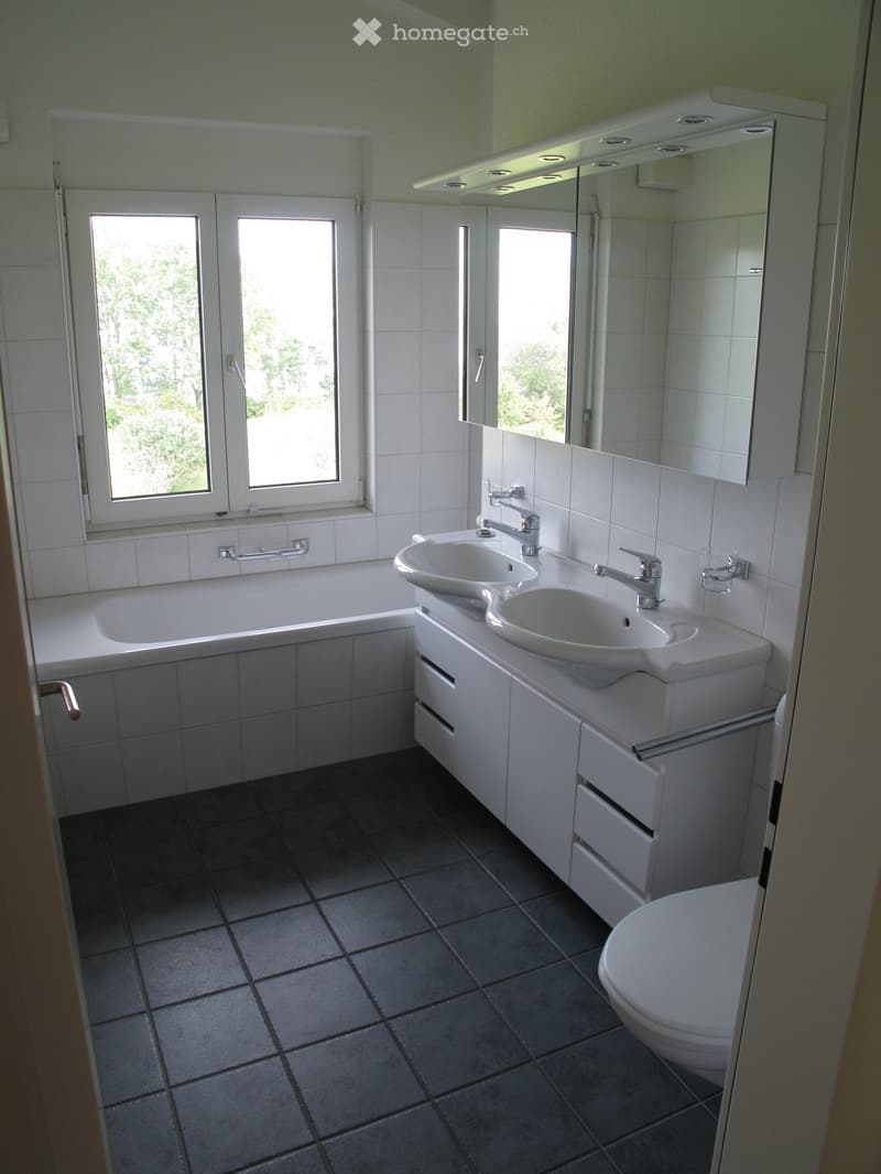 Modern bathroom with white tiles, a bathtub, double sink, mirror, and toilet. Features large windows with a view of greenery outside.