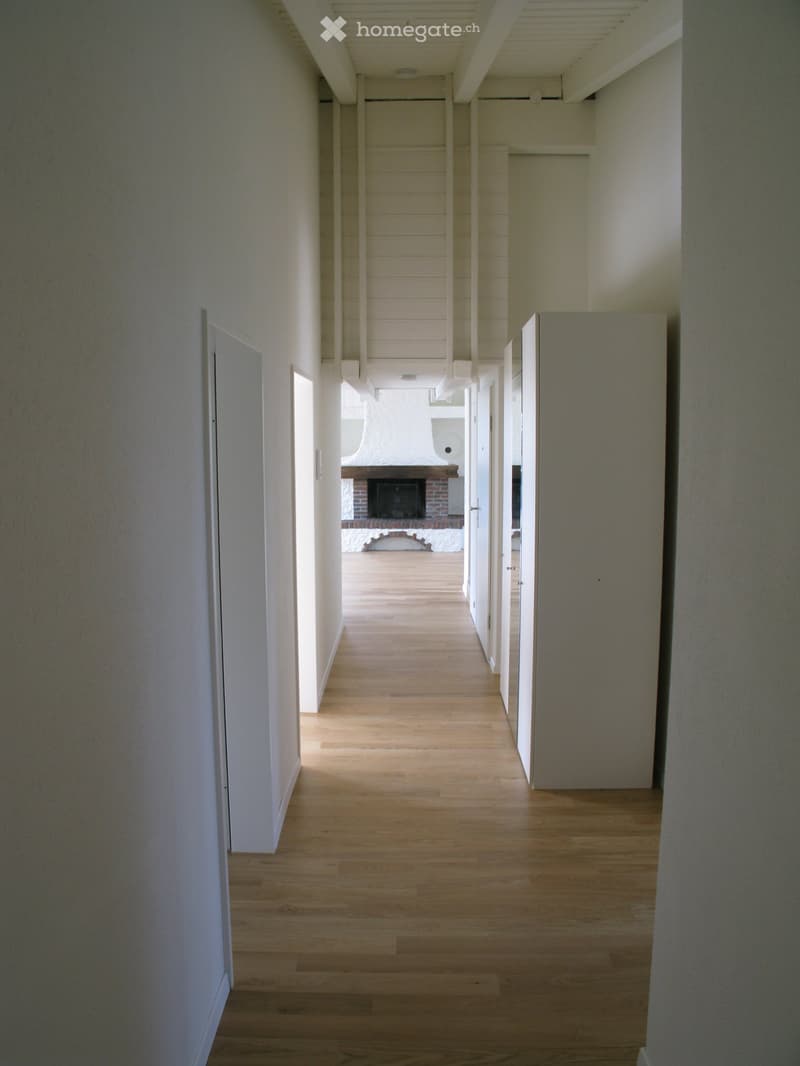 A long, narrow hallway with light wooden flooring and white walls, leading to a room with a fireplace.