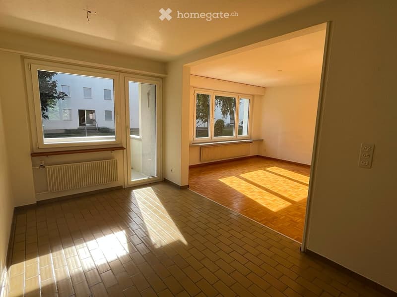 Empty room with tiled and wooden flooring, large windows, radiators, and a sliding glass door leading to another room.