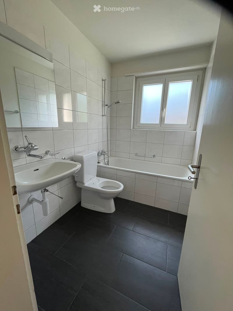 Modern bathroom with white tiles, black floor, sink, toilet, bathtub, and window with frosted glass.