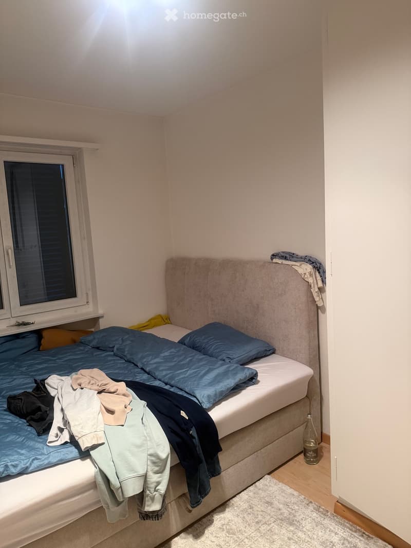 A small bedroom with a single bed, blue pillows, and a blanket. Clothes are draped over the bed, and a bottle is placed on the floor near the bed.