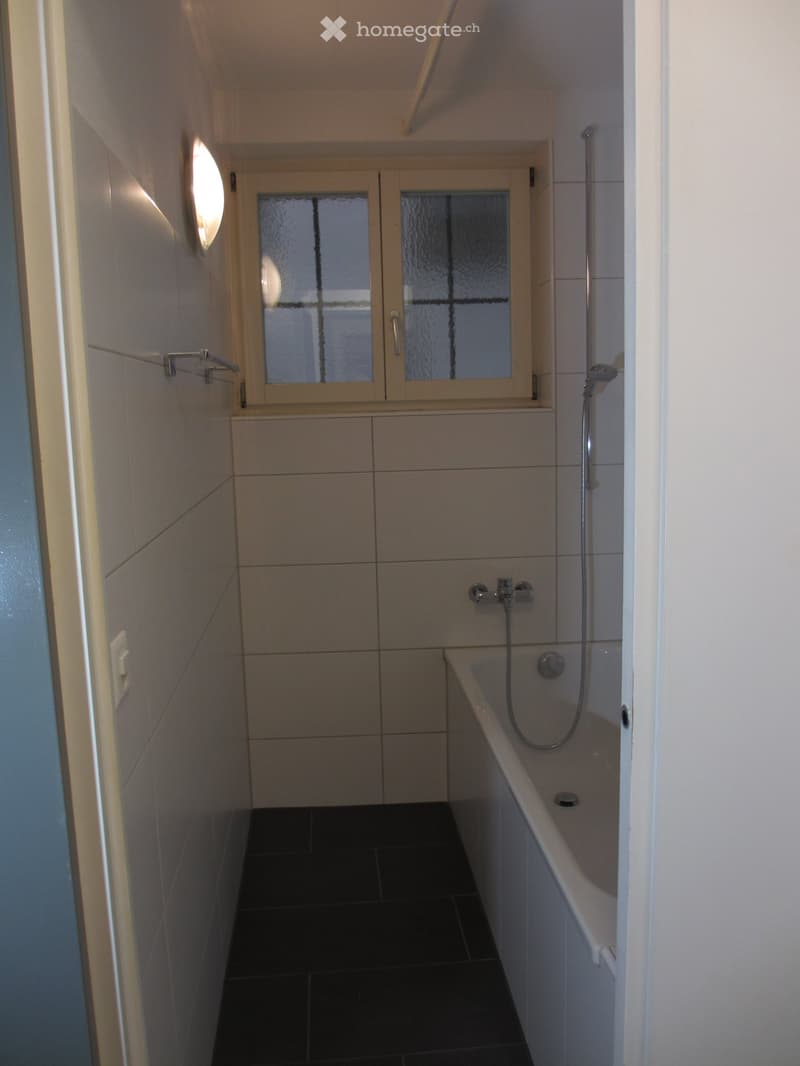 Modern bathroom with a bathtub, shower, and frosted glass window. Features dark tiled flooring and light-colored tiled walls.