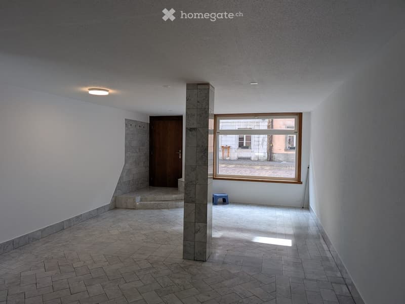 Empty hallway with tiled floor, white walls, a central column, a wooden door, and a large window showing an outdoor view.