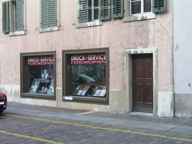 Facade of a two-story building with green shutters, large windows, and a storefront displaying signs for "DRUCK-SERVICE FOTOKOPIEN".