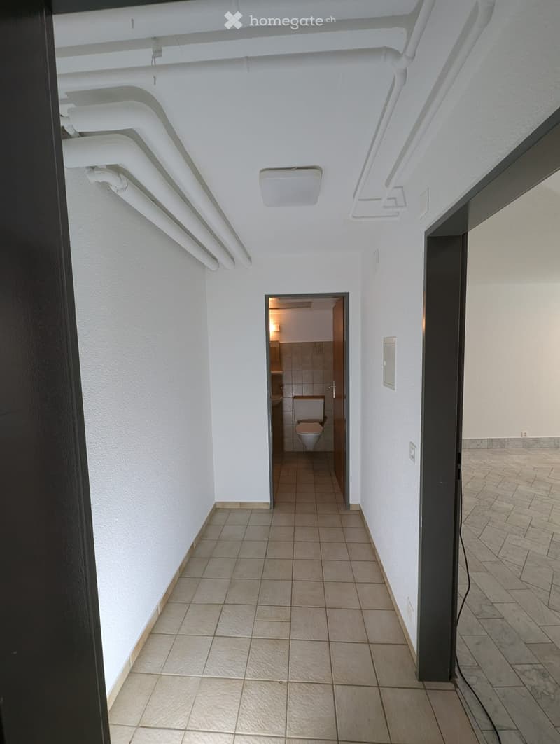 A clean, tiled hallway with white walls and a visible bathroom at the end, featuring a toilet and sink.