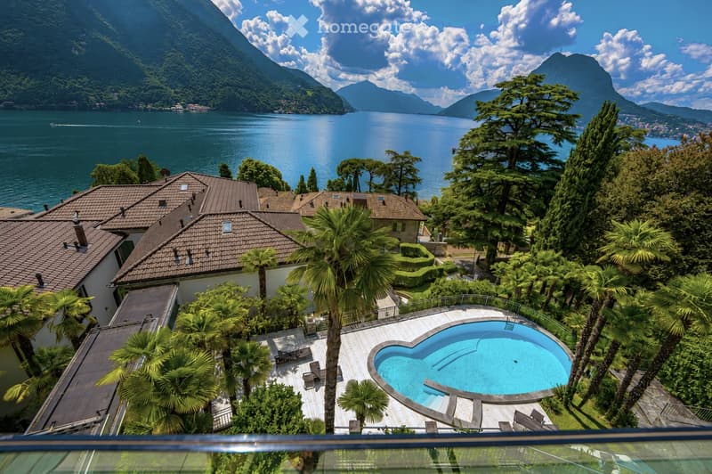 Aerial view of a luxurious villa with a heart-shaped swimming pool, surrounded by lush greenery and palm trees, overlooking a serene lake and mountain landscape.