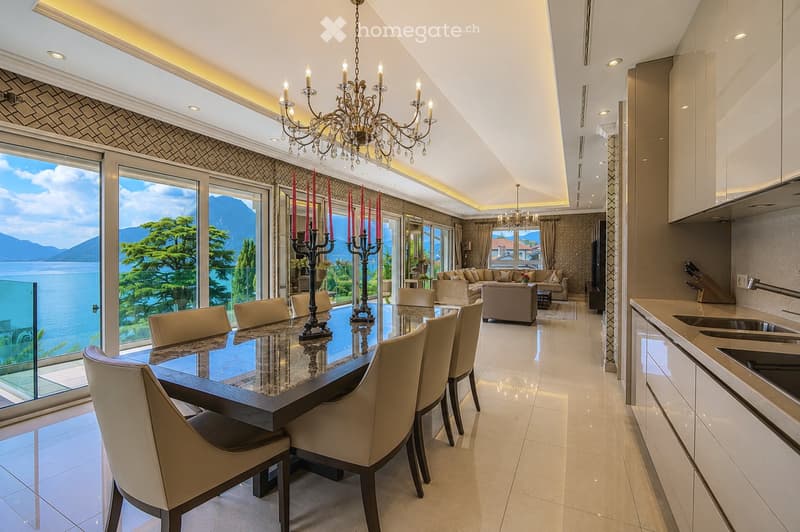 Elegant dining area with a large glass table, six chairs, and a candelabra centerpiece, connected to a modern kitchen with white cabinets and stainless steel appliances. The space features large windows offering a scenic view of a lake and mountains.