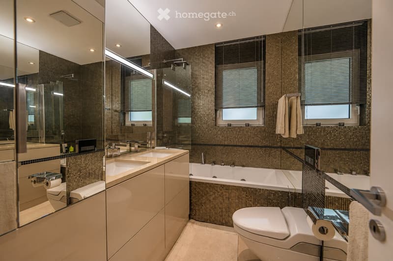 Modern bathroom with a large mirror, double sink vanity, bathtub, toilet, and towel racks. Features dark tiled walls and large windows with blinds.