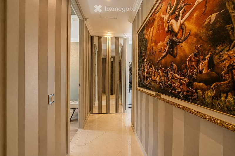 Elegant hallway with light-colored walls, polished tile flooring, recessed lighting, and a large framed painting on one wall. The hallway features a mirrored sliding door at the end, leading to another room.