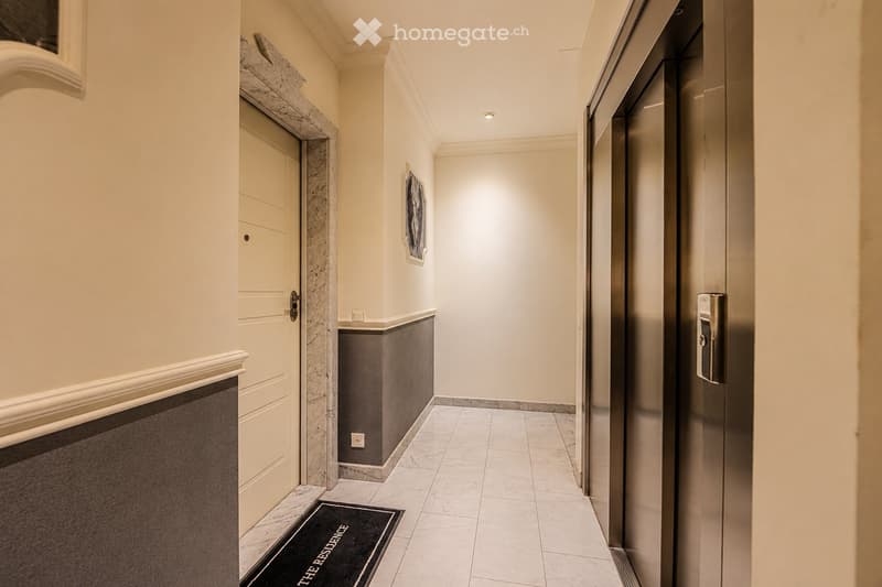 Elegant hallway with marble floors, white walls, and a closed elevator door on the right. A framed picture hangs on the wall, and a small black doormat with the text "THE RESIDENCE" is placed near the door on the left.