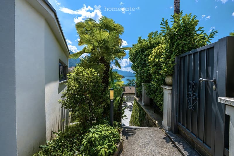 A narrow pathway lined with lush greenery and palm trees leads to a gated entrance, with a view of the sea and mountains in the background.