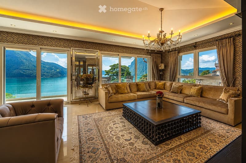 Elegant living room with large windows offering a scenic mountain and lake view, featuring a beige sectional sofa, dark wooden coffee table, armchair, chandelier, and decorative mirrors.