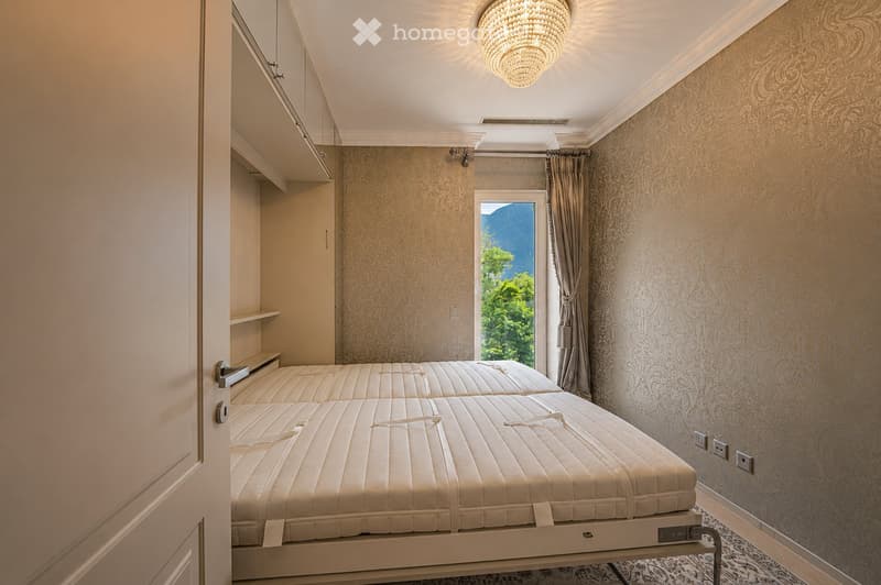 Elegant bedroom with two single beds, patterned wallpaper, large window with scenic mountain view, chandelier, and built-in cabinetry.