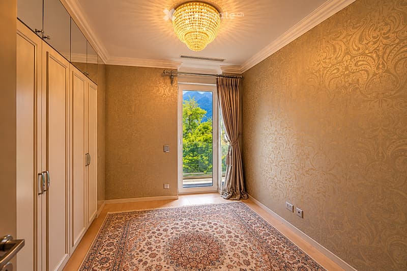 An empty room with patterned wallpaper, wooden flooring, a large window with curtains, and a chandelier on the ceiling.