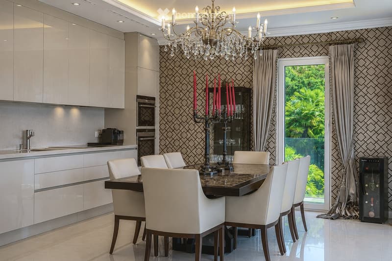 Modern kitchen and dining area with white cabinets, a dark dining table surrounded by white chairs, a chandelier, and a view of greenery through large windows.