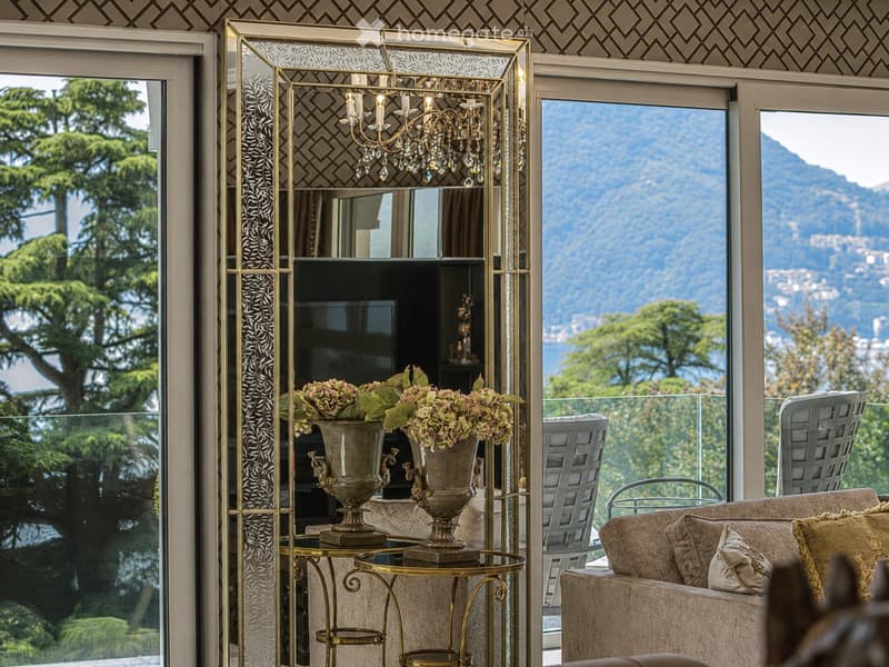 Elegant living room with a view of mountains, featuring a patterned sofa, gold side tables, a large vase with flowers, and a chandelier.